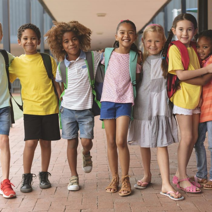 Front view of happy school kids standing in corridor and holding together in arms